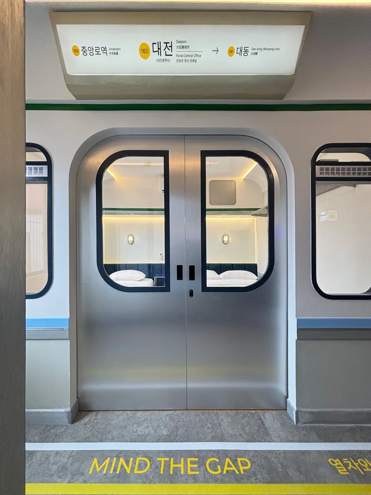 Dae Jeon: Pintu ala kereta bawah tanah dengan tulisan Mind the Gap, detail kecil bertema stasiun.